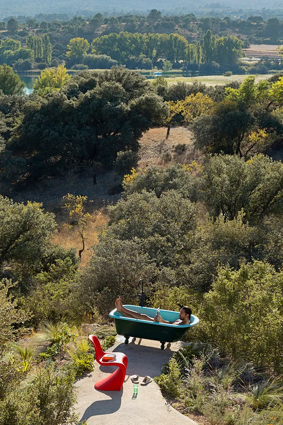 Mann entspannt in freistehender Badewanne auf Terrasse mit Blick über Olivenhain, See und roten Designstuhl.
