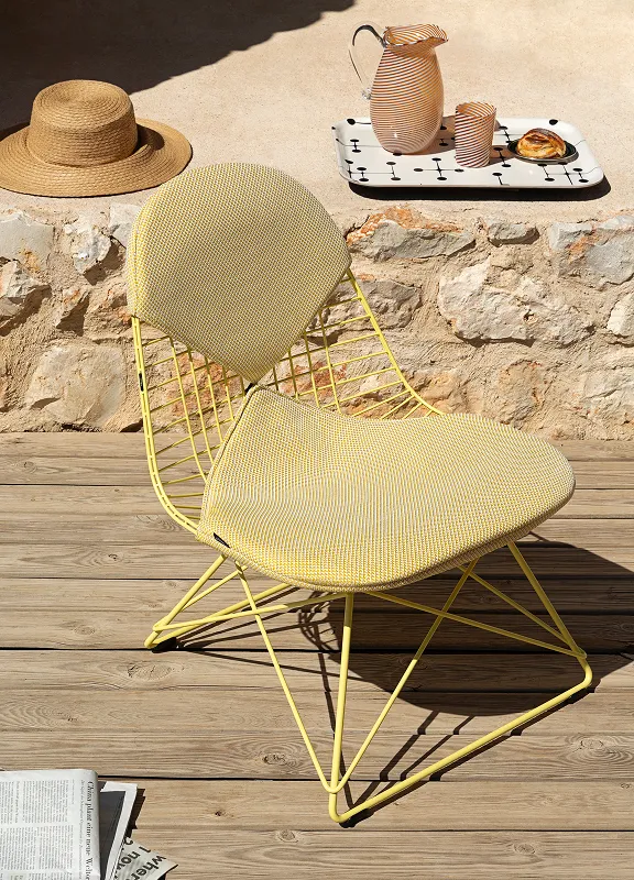 Gelber Wire Chair auf Holzterrasse vor Steinmauer, daneben Tablett mit Karaffe, Glas und Gebäck.